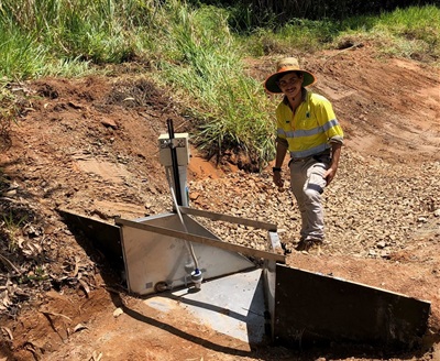 Worker in high-vis gear beside monitoring device in a dry, rocky drainage area.